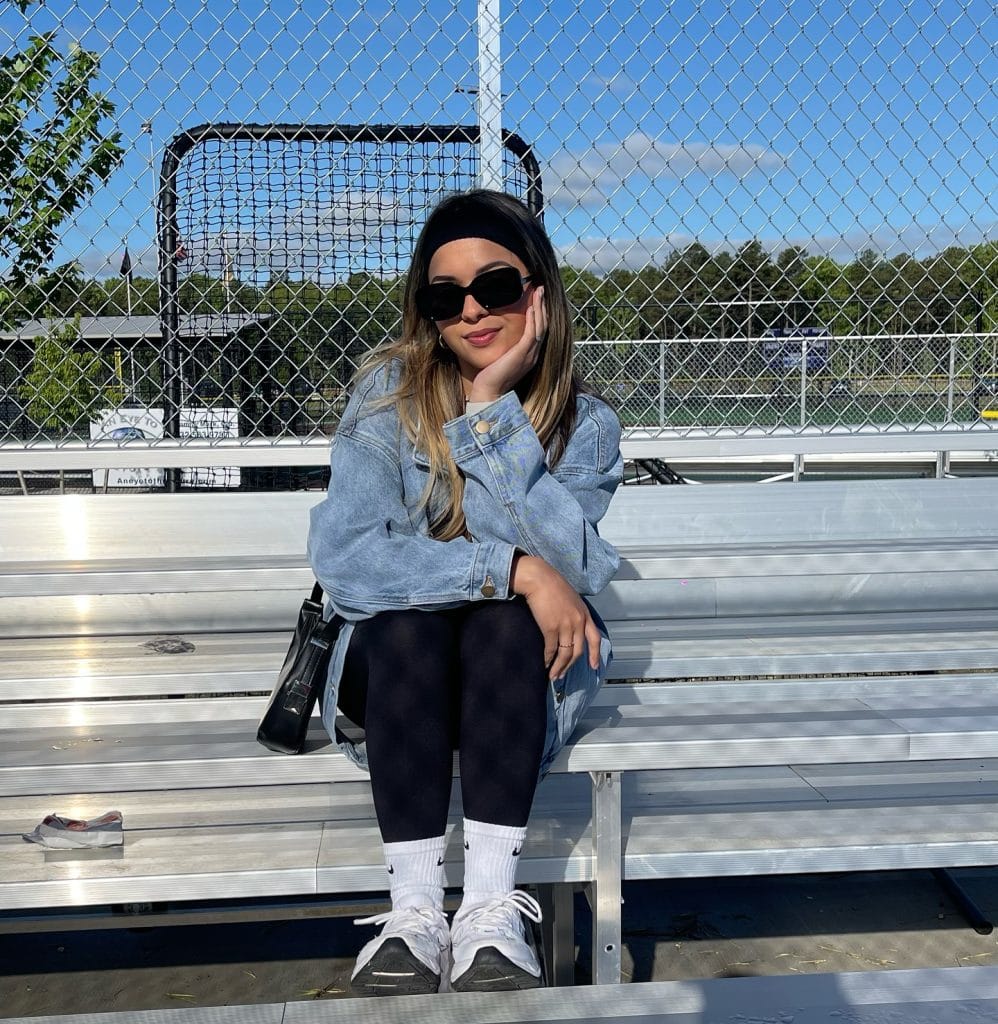 Person sitting on bleachers in sunglasses and denim jacket.