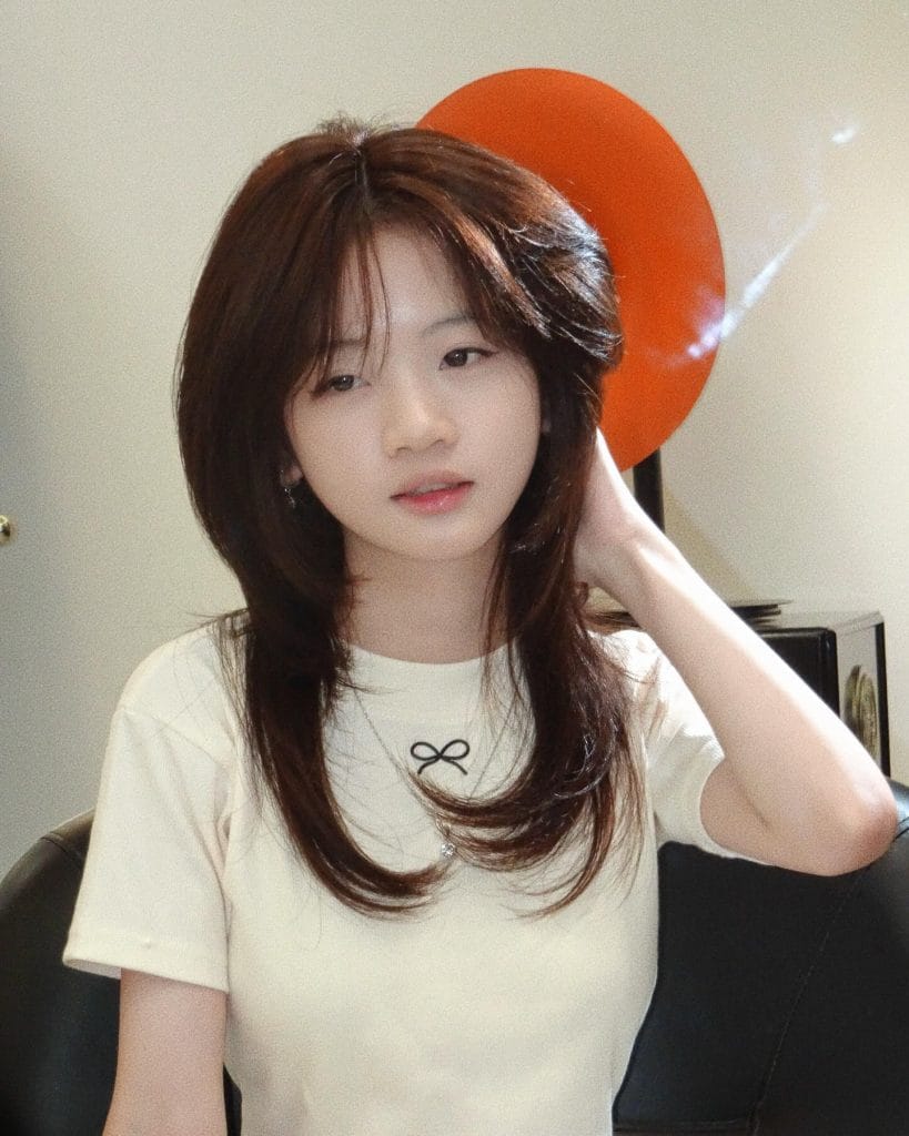 Person with long hair sitting indoors, thoughtful expression.