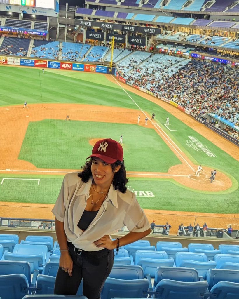 Smiling woman in baseball stadium with cap on.